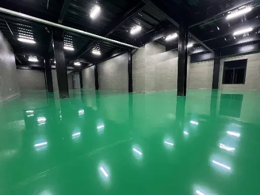 The image depicts a large warehouse space with a black ceiling and columns, grey walls, and green epoxy flooring by Carmacoat 