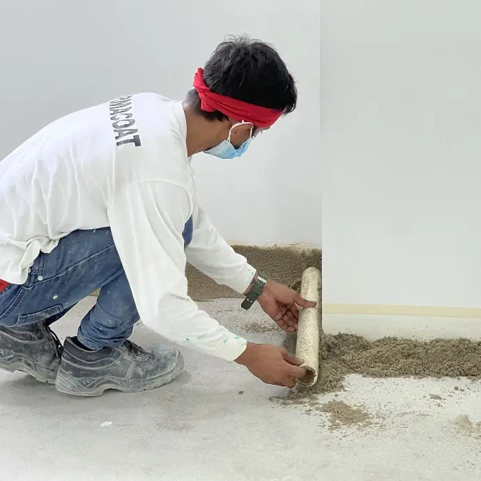 The image depicts a Carmacoat employee  installing floor coving by holding a PVC pipe horizontally against the corner where the floor and wall meet, using it to push an epoxy sand mix into the corner to create a quarter C cove